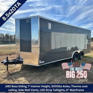 Enclosed dual‑axle cargo trailer in gray on a dirt lot with sale graphics and an American flag banner.