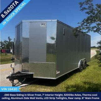Silver enclosed cargo trailer with dual axles, parked on grass; blue '8.5x20TA IN-STOCK SPECIAL' banner across corner and VIN 16430 visible.
